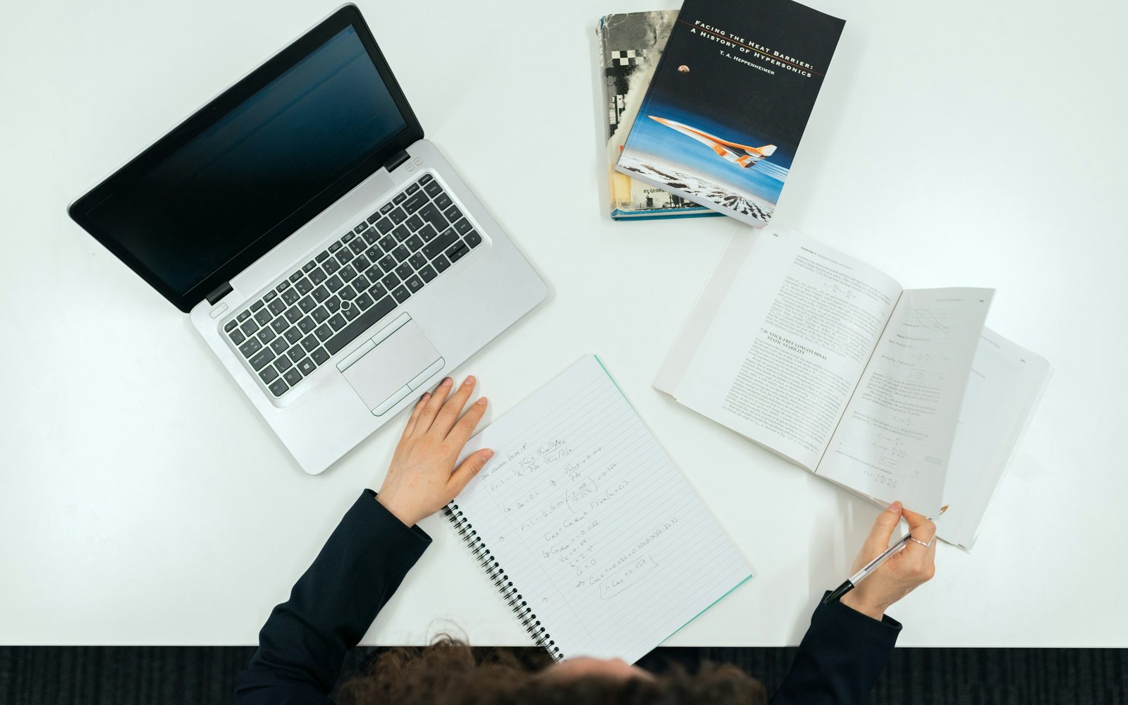 Person studies at a white desk with an open laptop, notebook, and reference books.