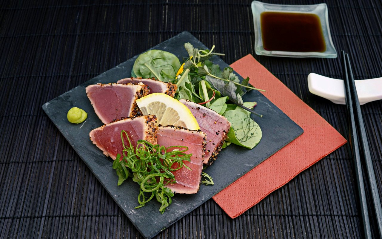 Pink-centered seared tuna slices with lemon wedges, mixed greens, scallions, and wasabi on a black slate plate.