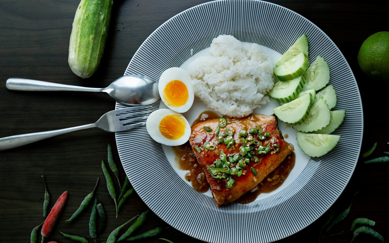 Glazed cooked salmon with white rice, cucumber slices, two egg halves, and chopped green chile on a striped plate.