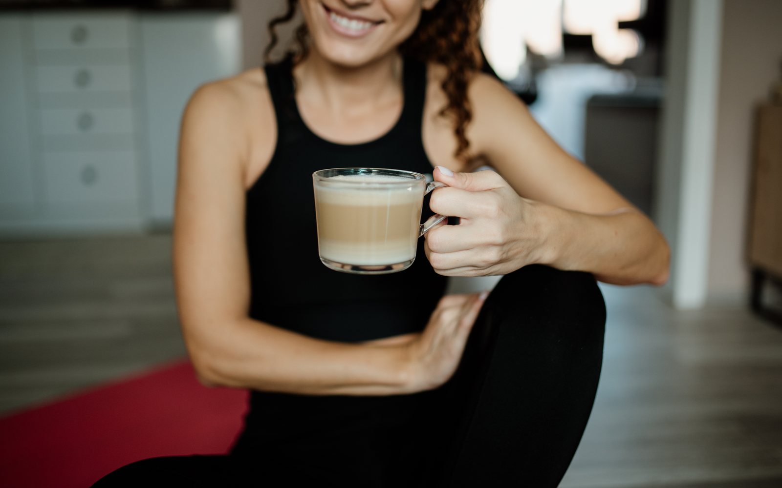 Smiling woman in workout clothes sits on a red mat holding a cup of coffee.