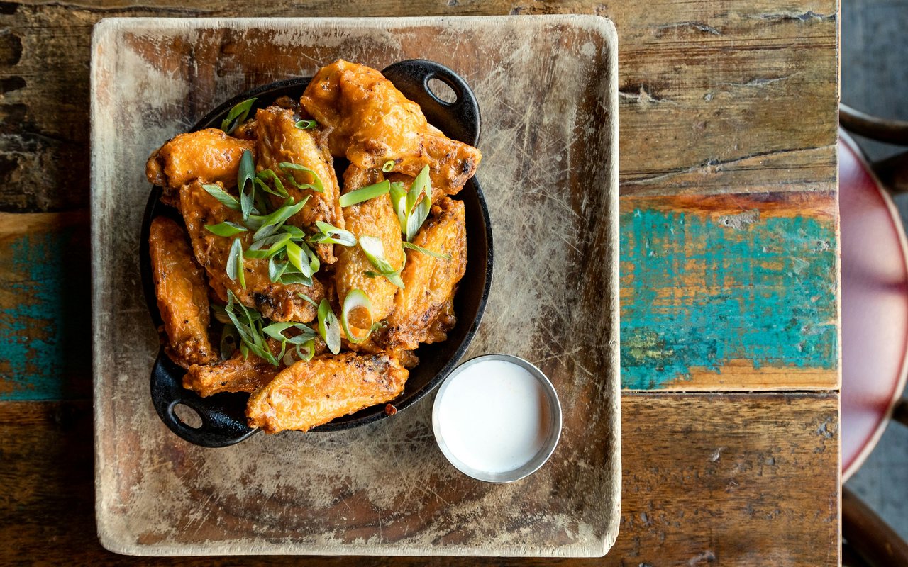 Buffalo-style chicken wings topped with scallions in a black skillet beside a cup of creamy dip.