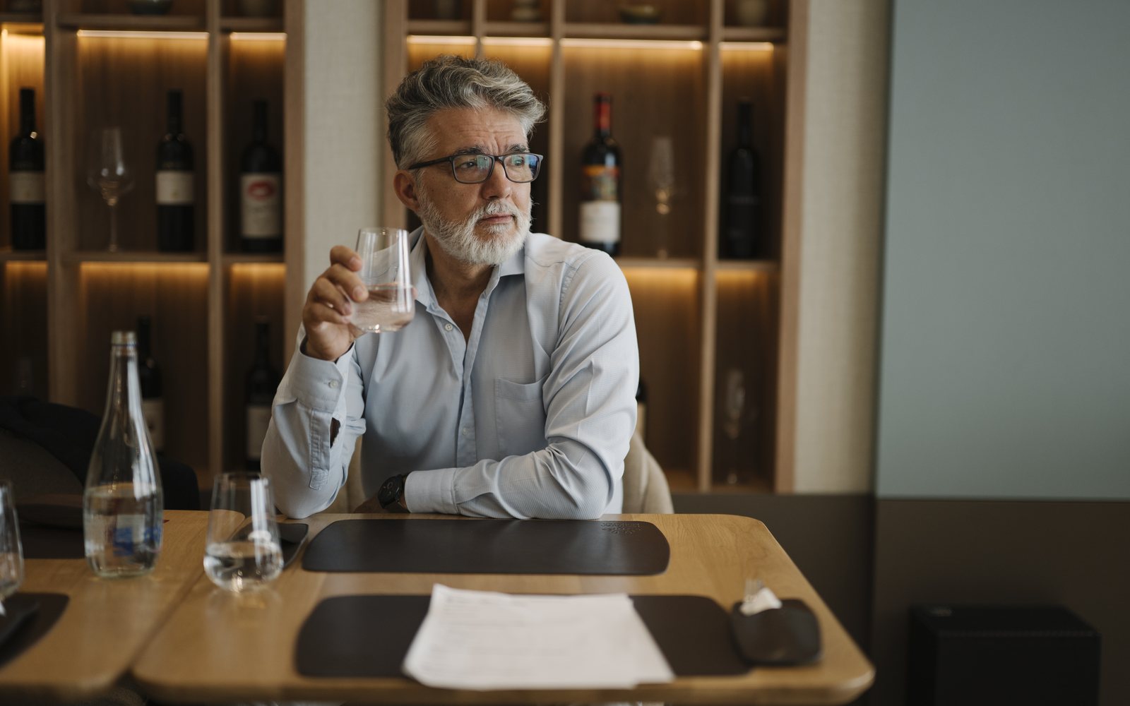 Man holding a glass of water at a table with wine bottles displayed behind him.