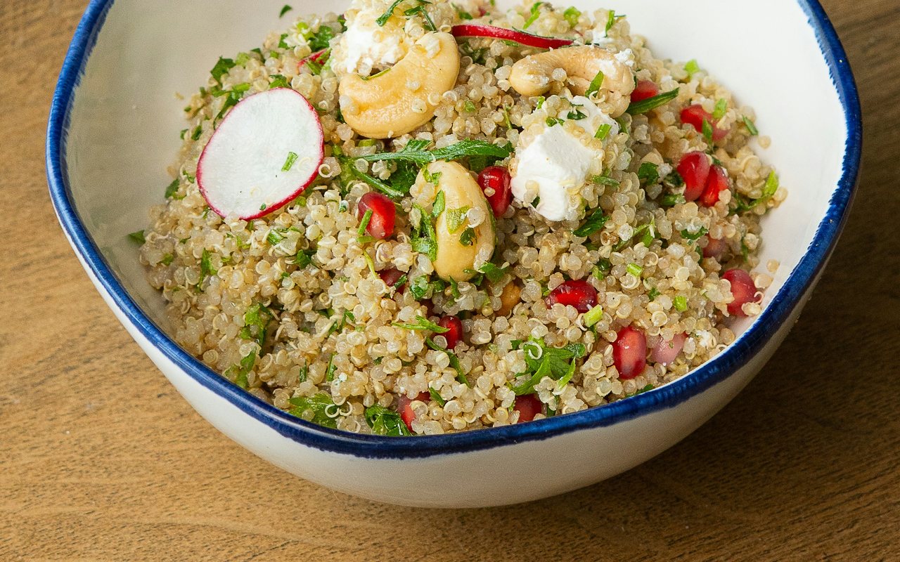 A ceramic bowl of quinoa salad with pomegranate seeds, feta, cashews, radish slices, and chopped herbs.
