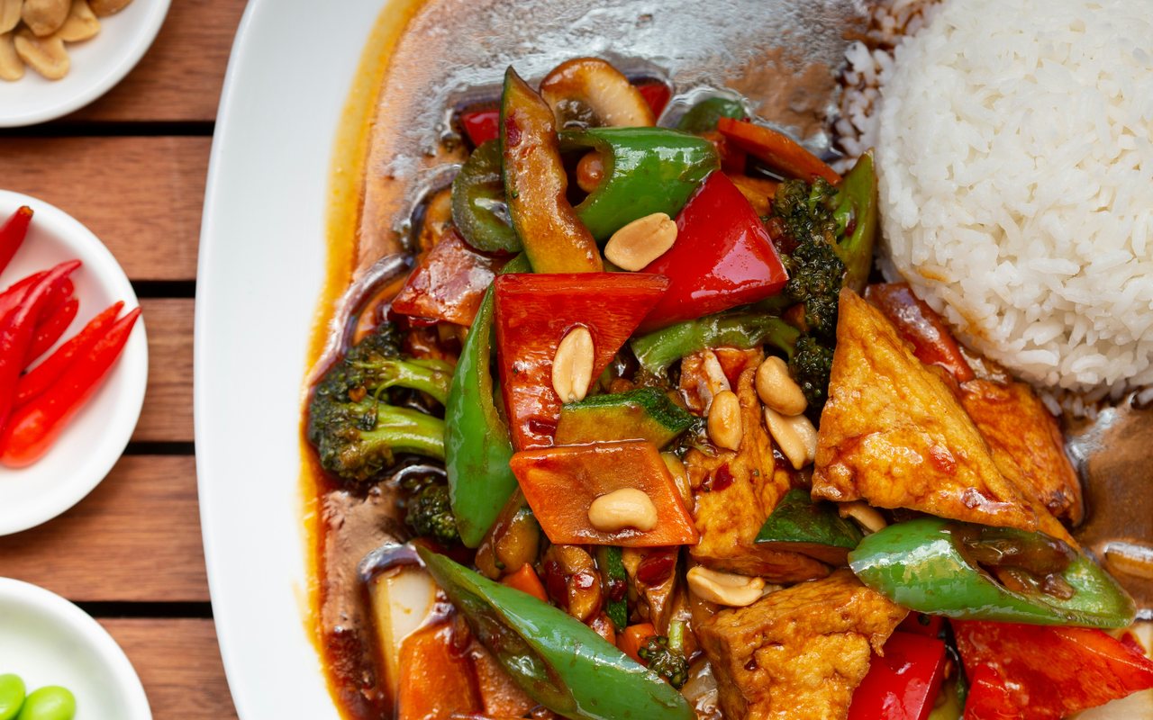 Sauced tofu with broccoli, bell peppers, mushrooms, peanuts, snap peas, and white rice on a white plate.