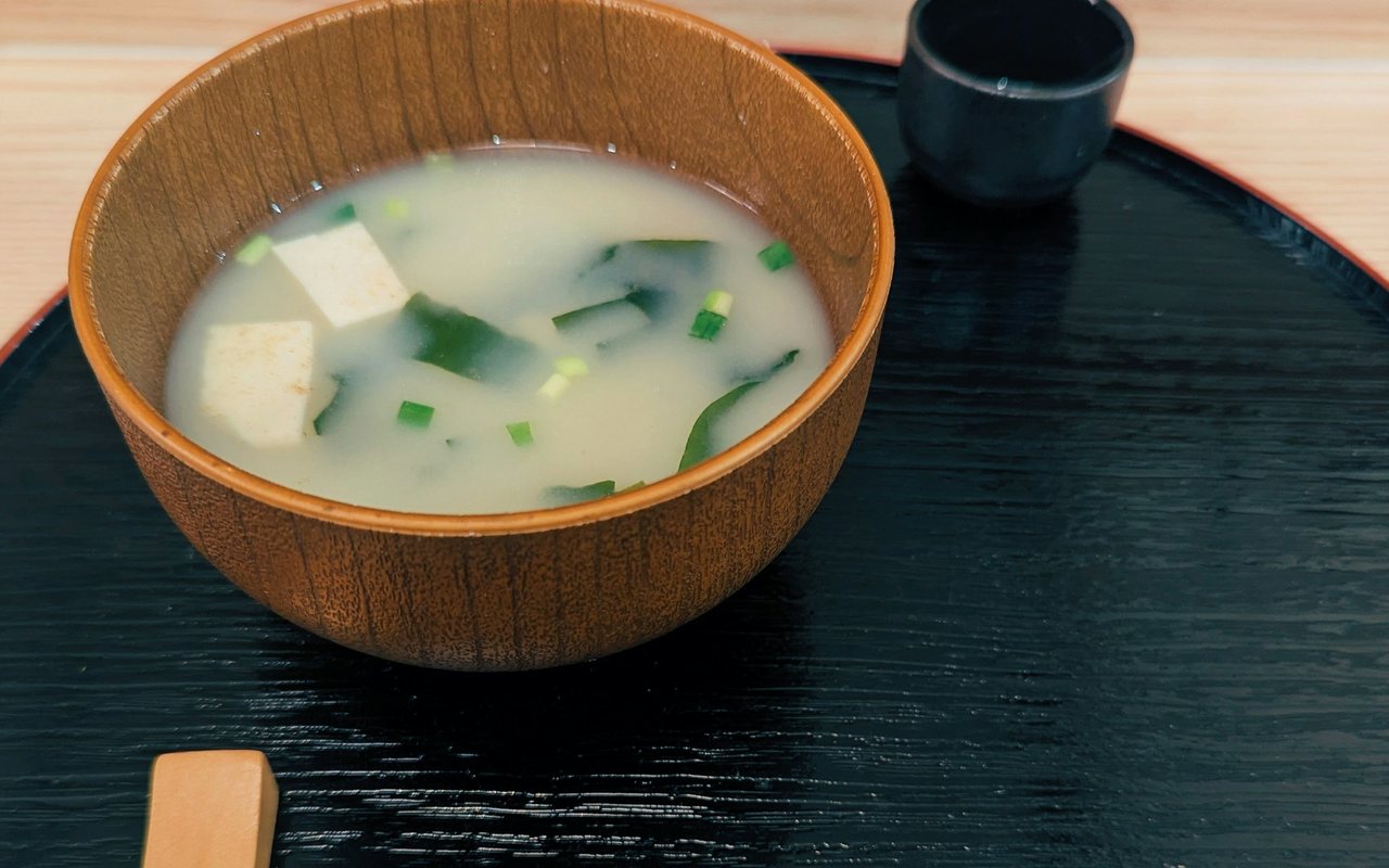 A wooden bowl of pale miso soup with tofu cubes, seaweed, and scallions on a black tray.