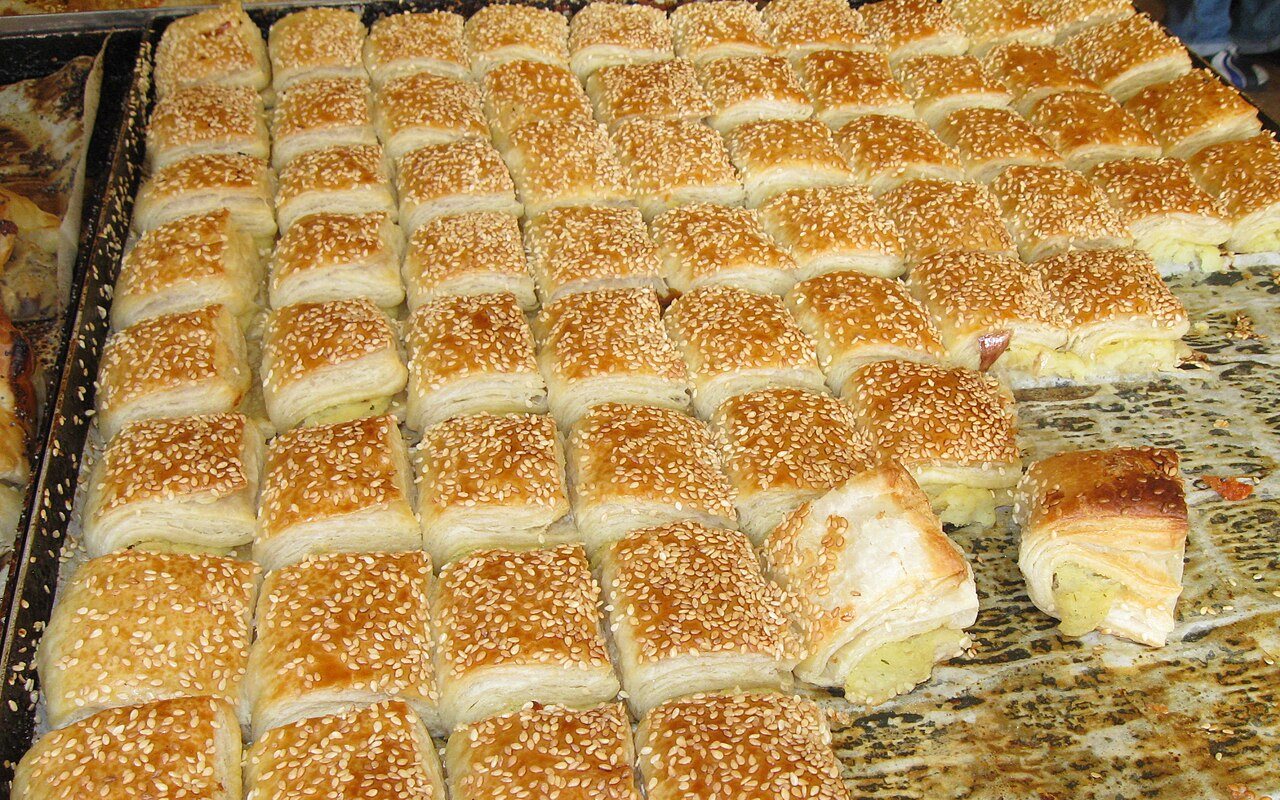 A tray of square sesame-topped börek, with two pastries cut open to show pale potato filling