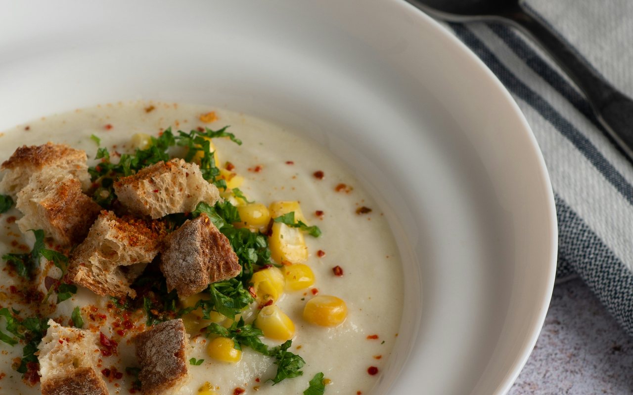 Creamy cauliflower soup puree topped with corn, herbs, red pepper flakes, and croutons in a white bowl.