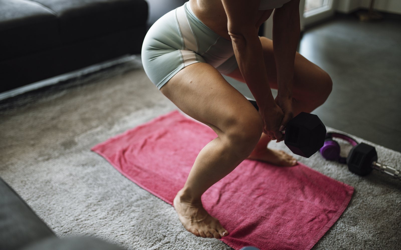 Adult holds a black dumbbell in a deep barefoot squat on a pink mat.