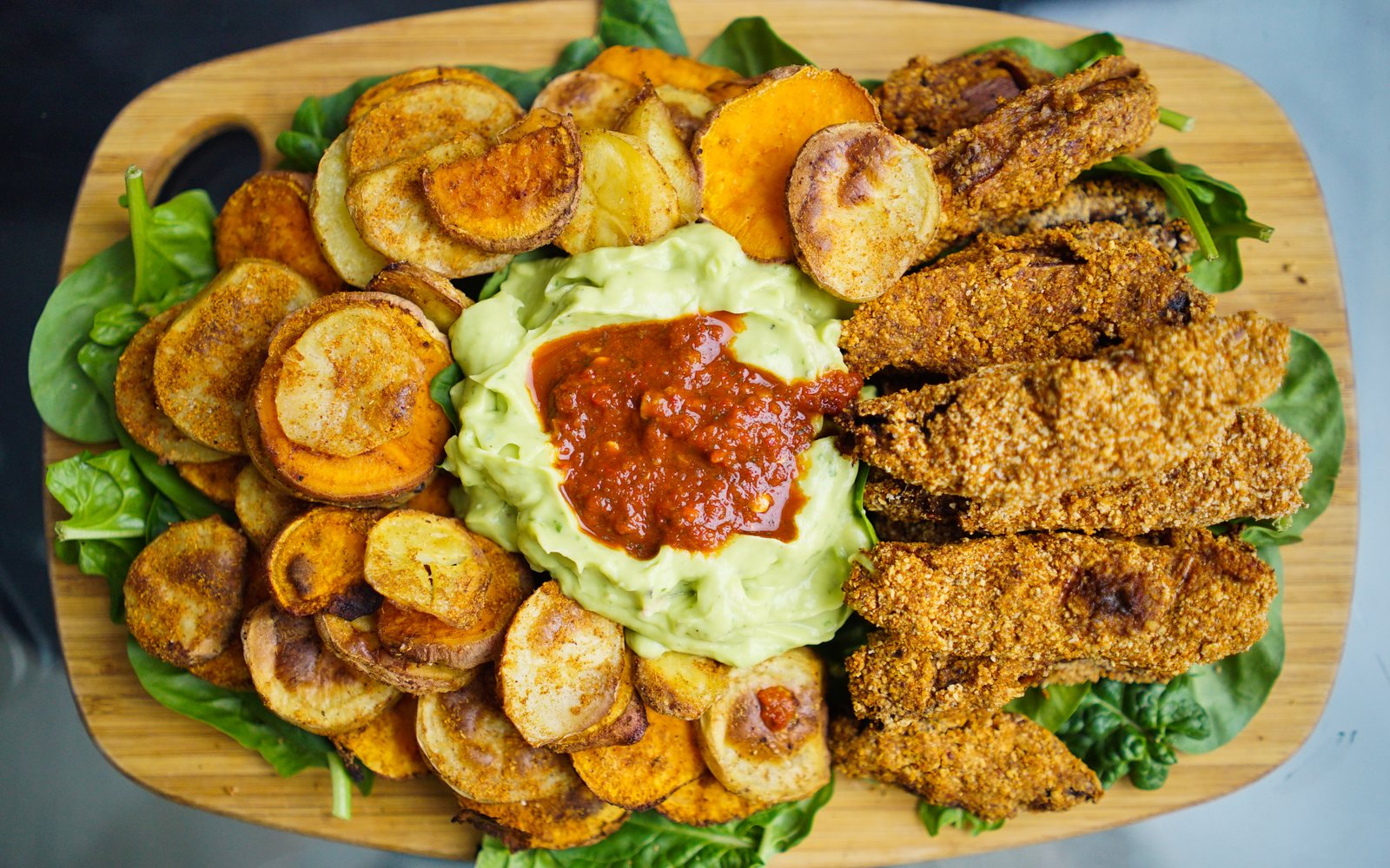 Platter of roasted potatoes, breaded strips, leafy greens, avocado dip, and red salsa.