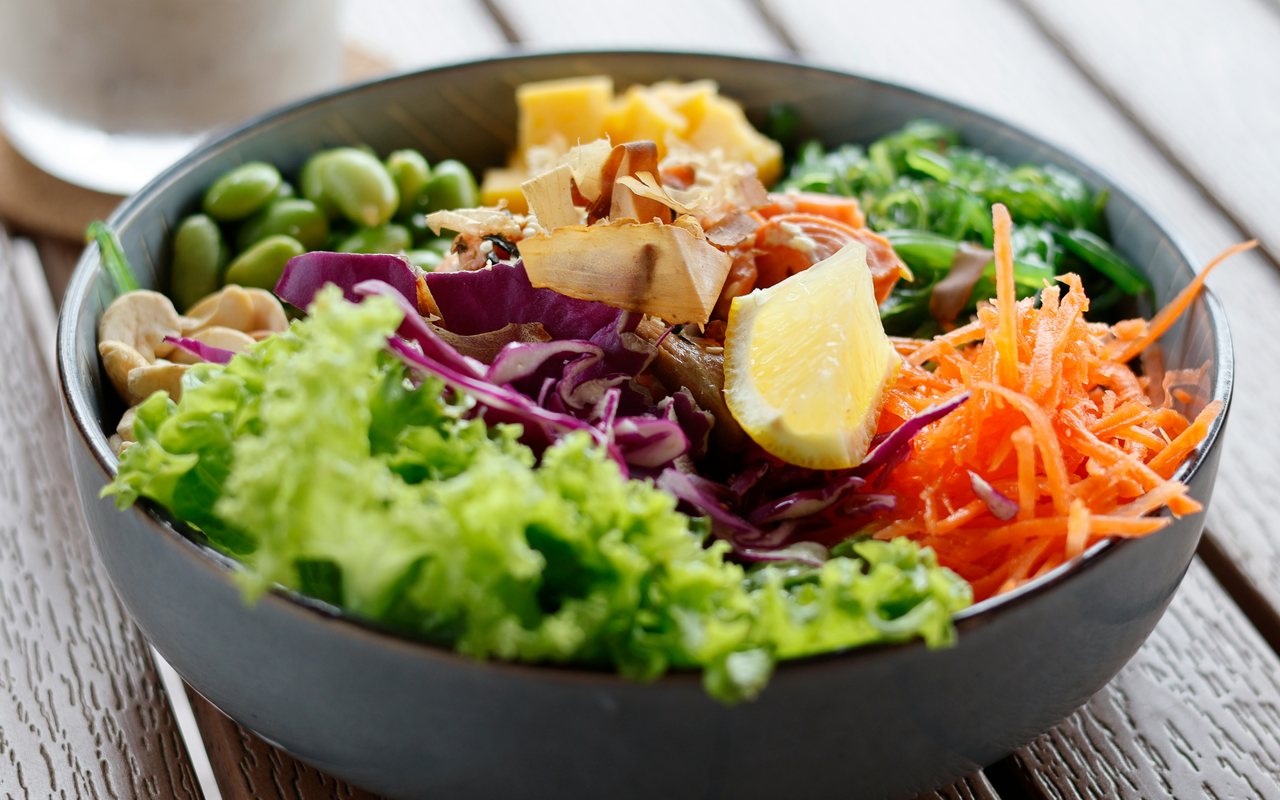 A salad bowl with shelled edamame, shredded red cabbage, carrots, lettuce, seaweed, lemon, and mixed toppings.