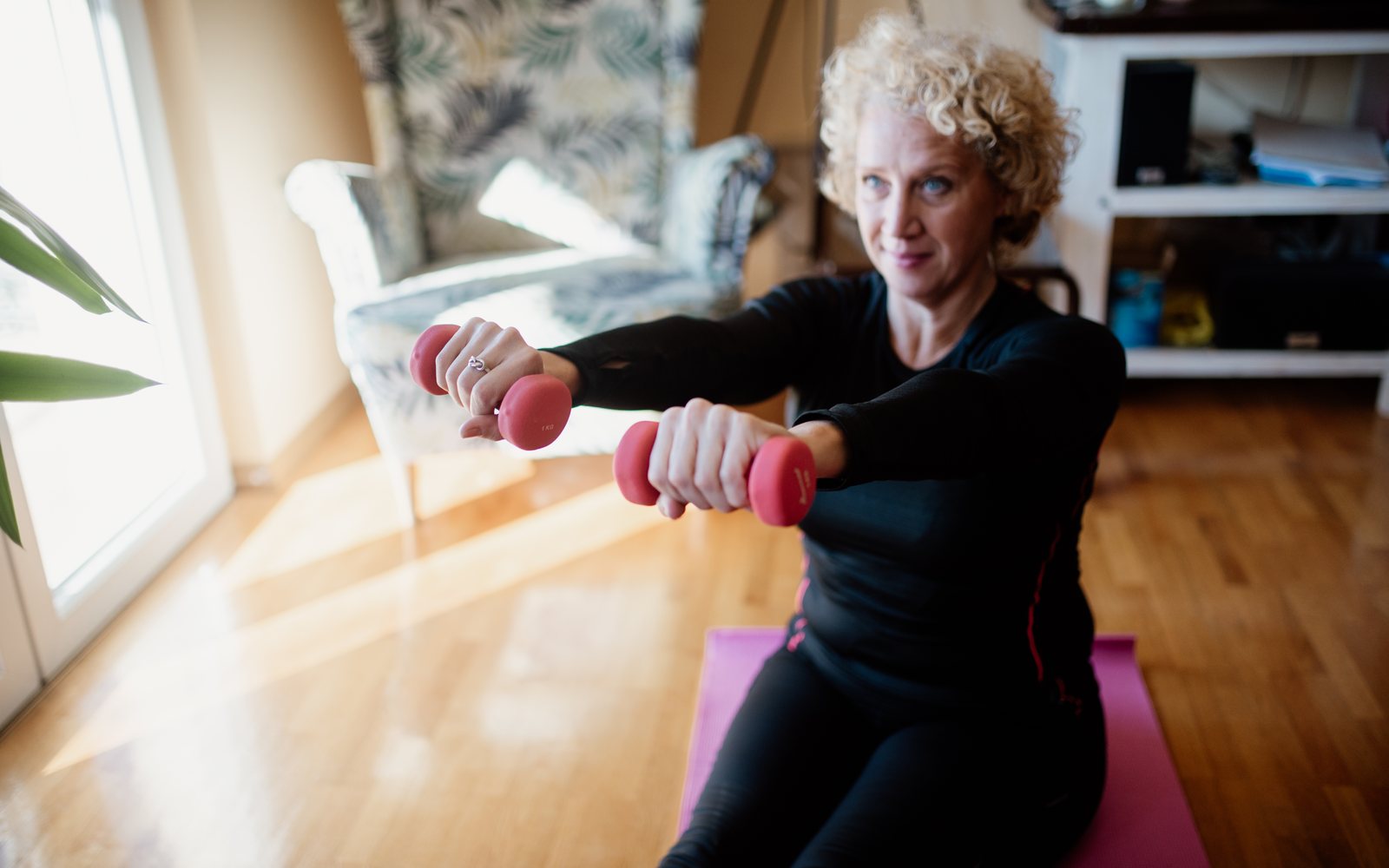 Older woman lifts a pair of red dumbbells in a bright living room.