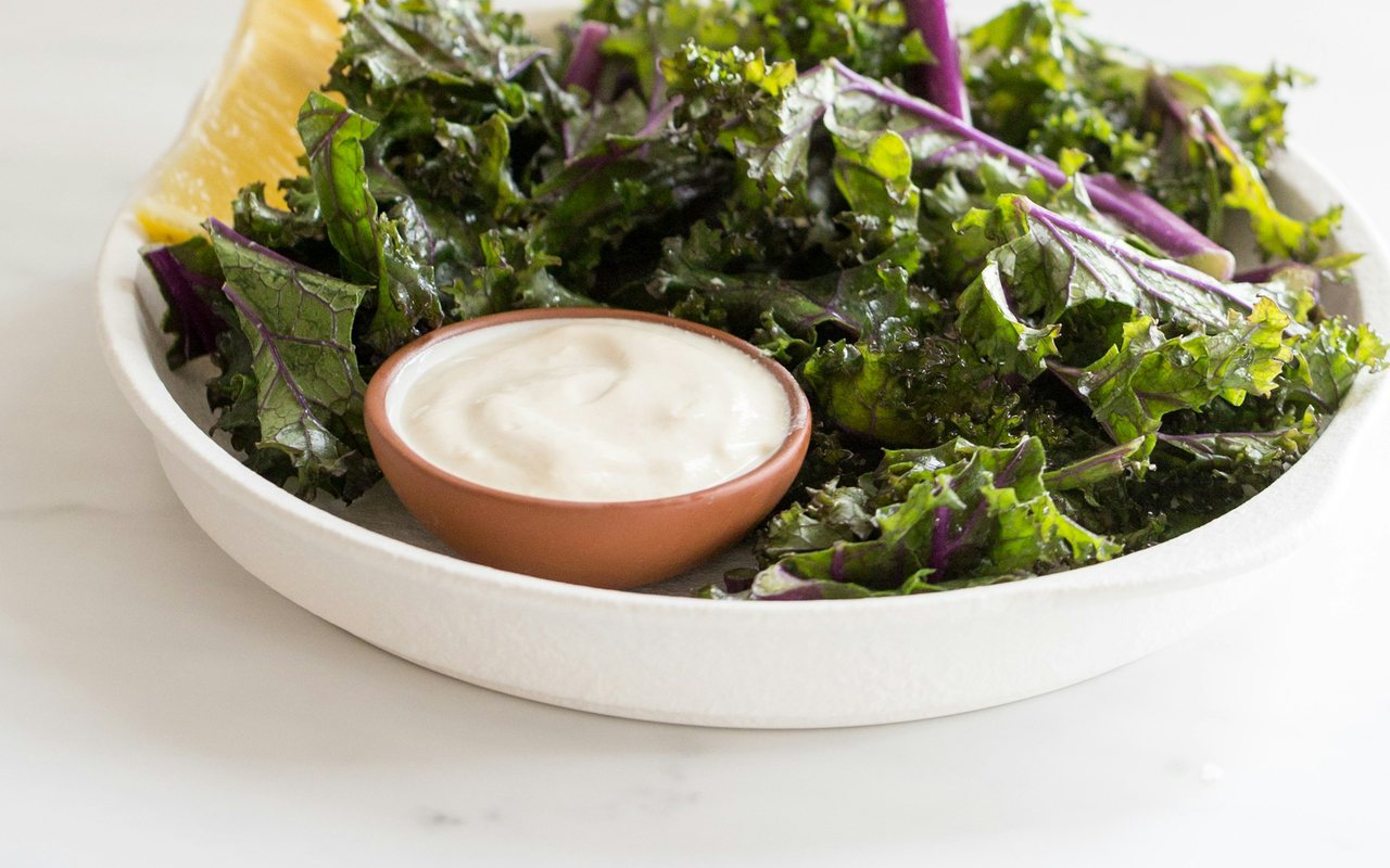 A white bowl of kale salad with purple stems, a lemon wedge, and a small bowl of tahini dressing.