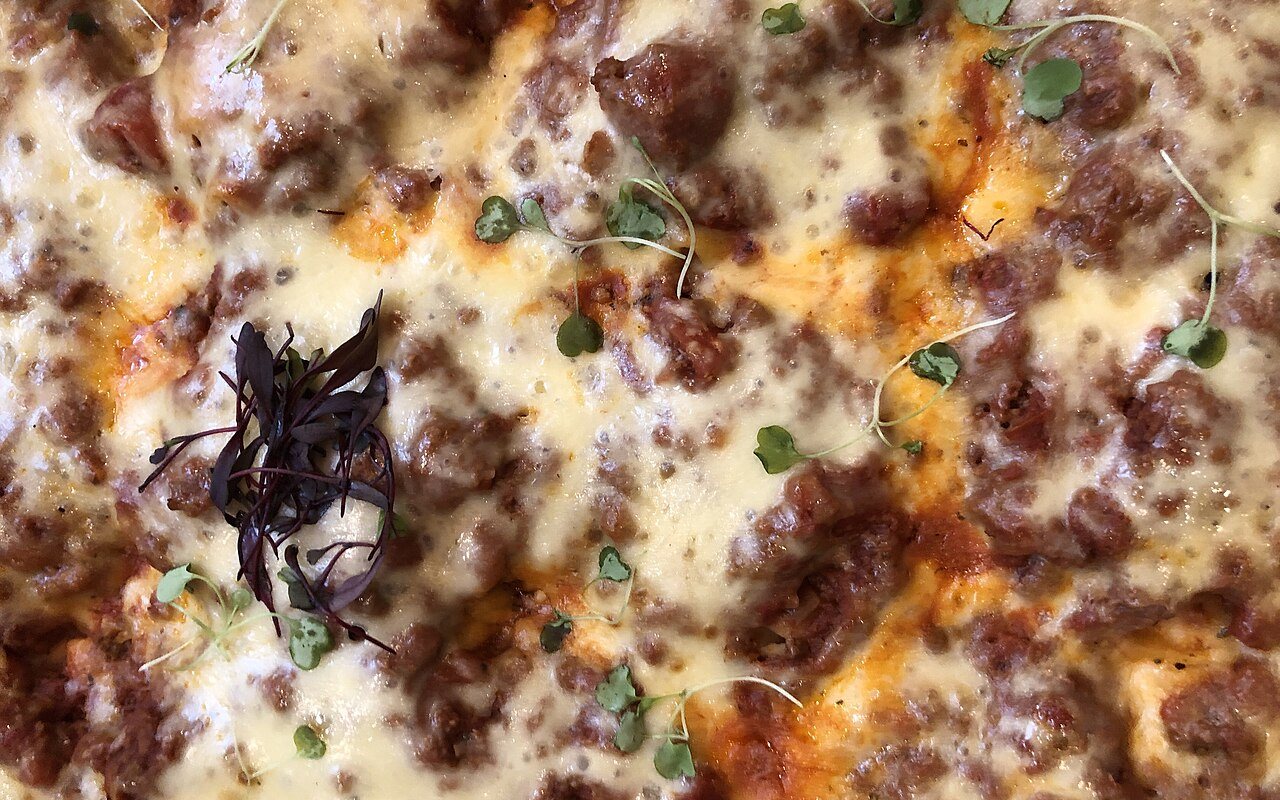 A square slice of beef lasagna with visible pasta, meat sauce, béchamel, and browned cheese layers