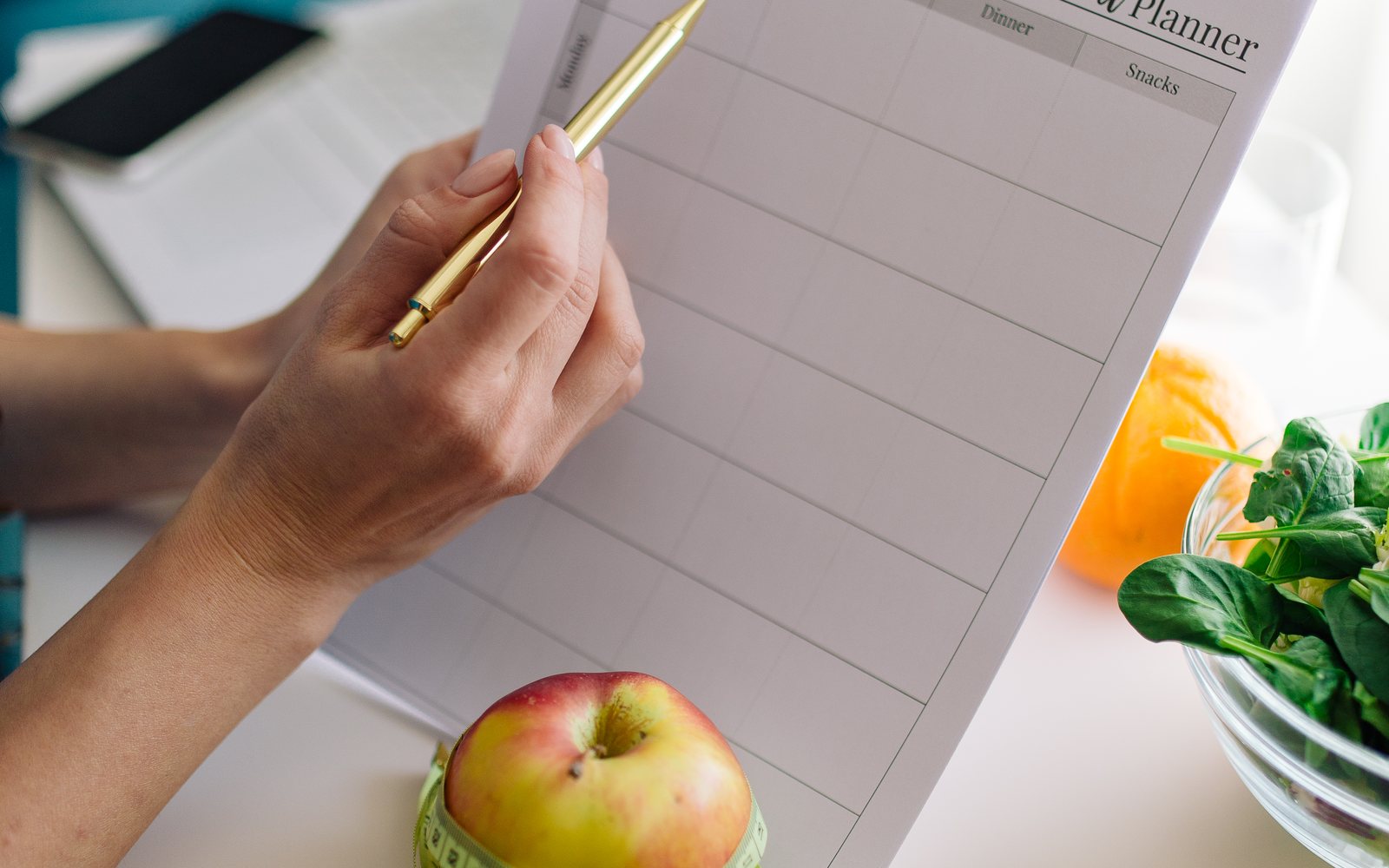 Person writes in a meal planner beside an apple and a bowl of spinach.