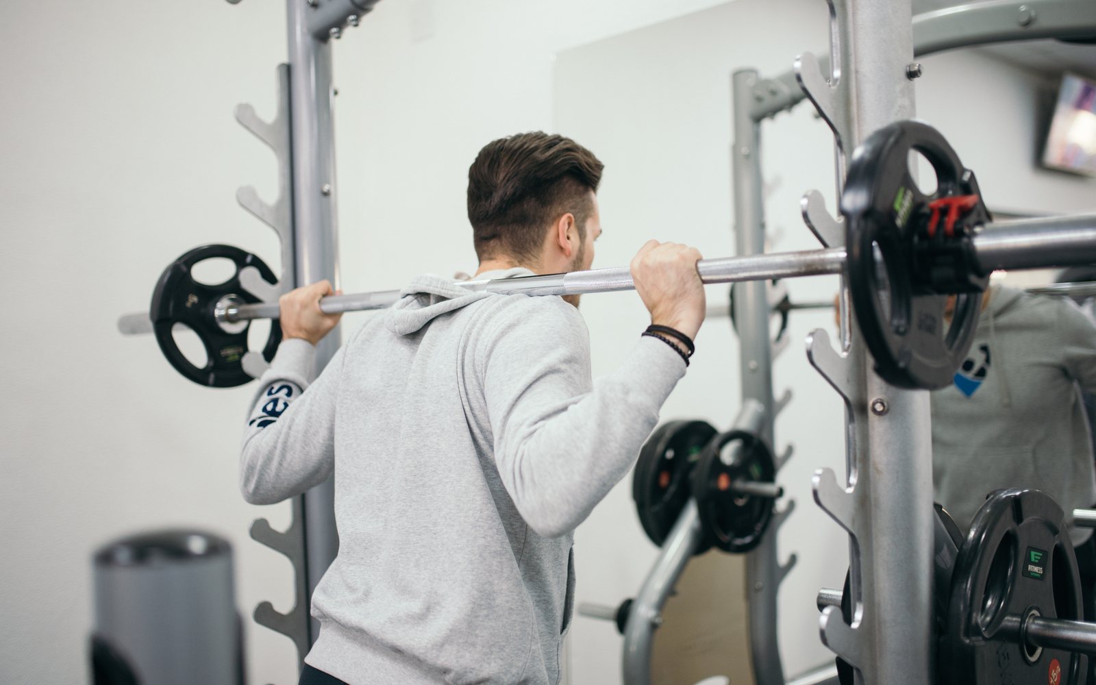 Man holds a barbell across his upper back inside a gym rack.