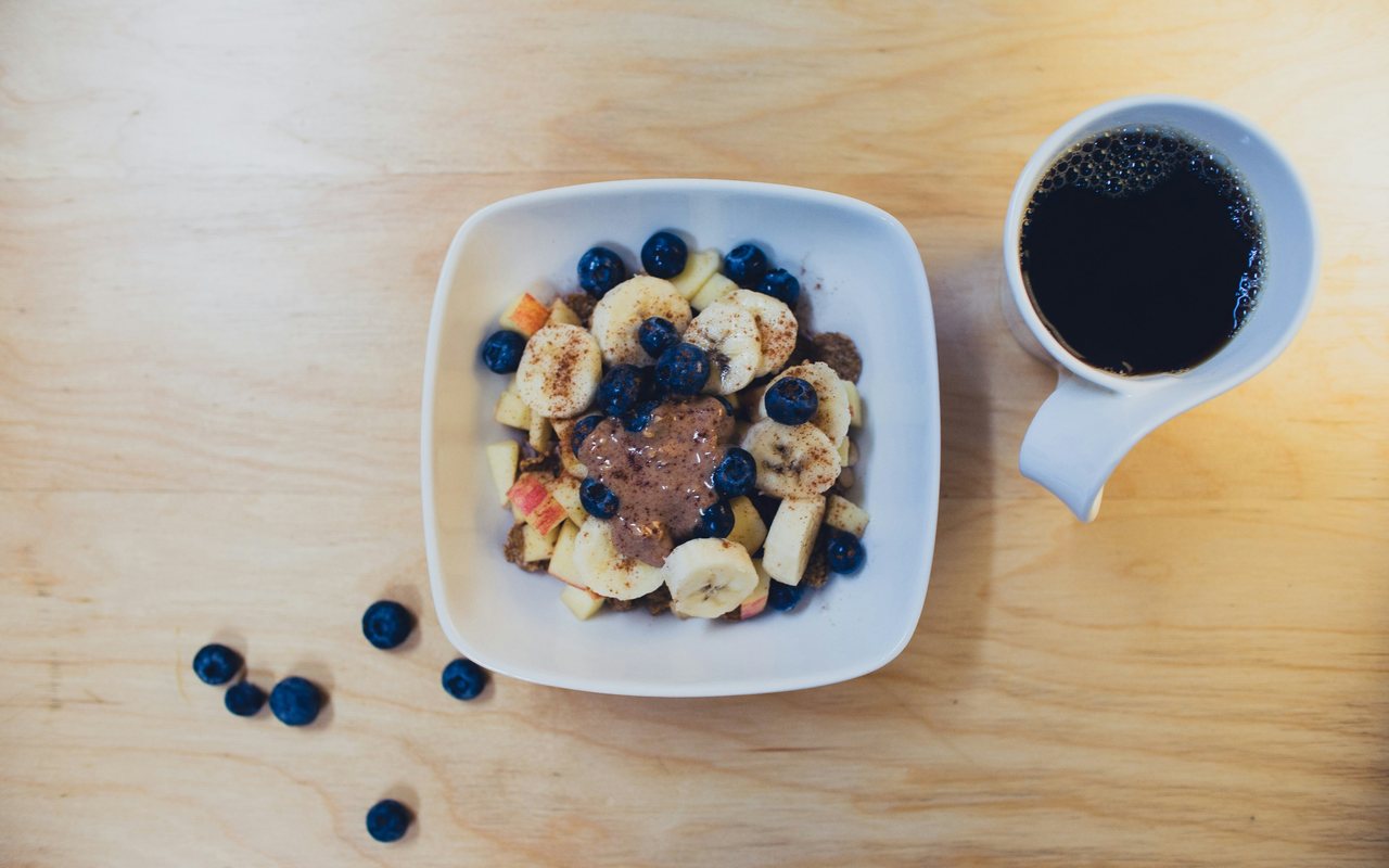 A square bowl of blueberry and banana oats topped with a visible peanut butter dollop on a pale wood table.