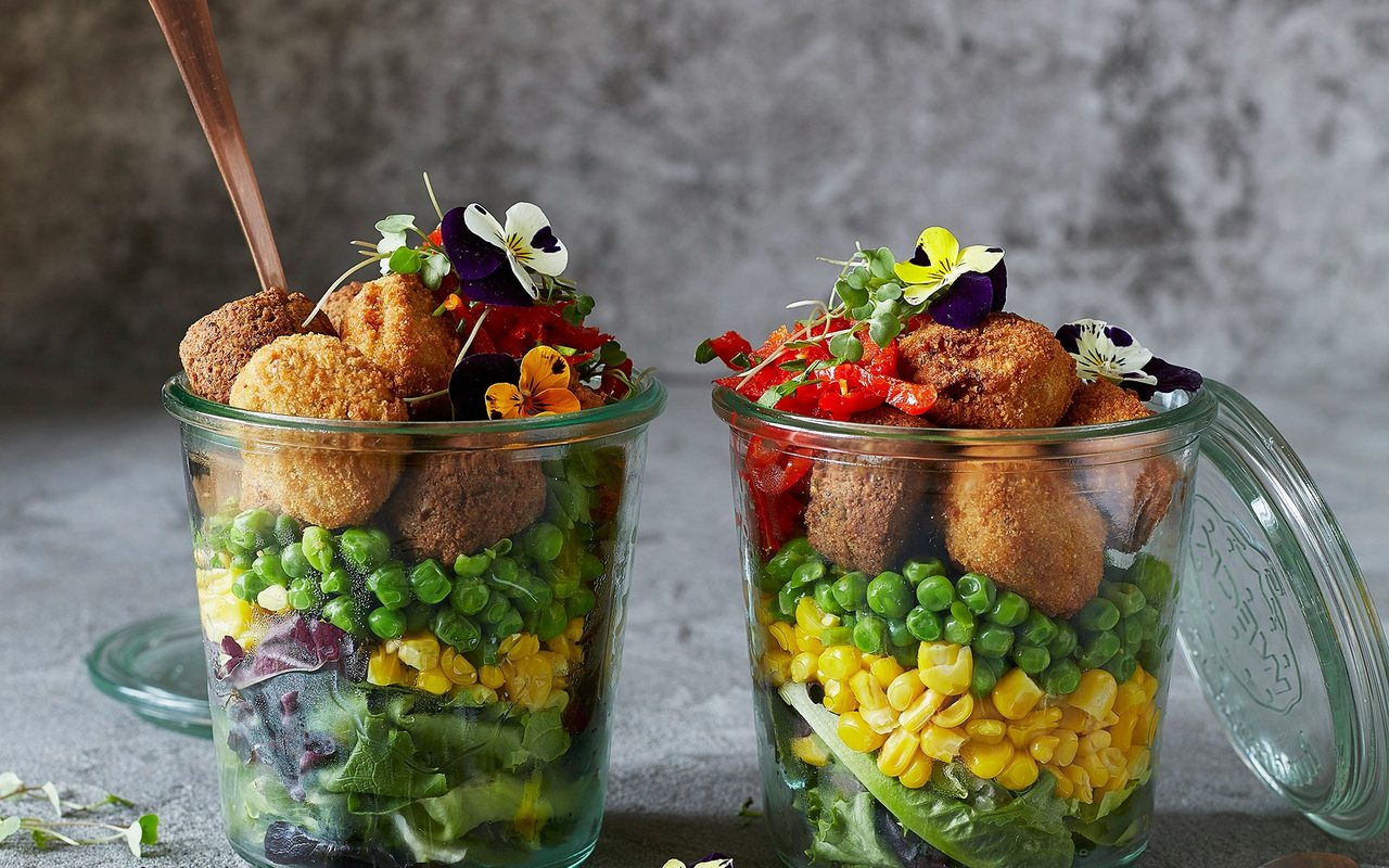 Two glass jars layered with lettuce, red cabbage, corn, green peas, peppers, and falafel.